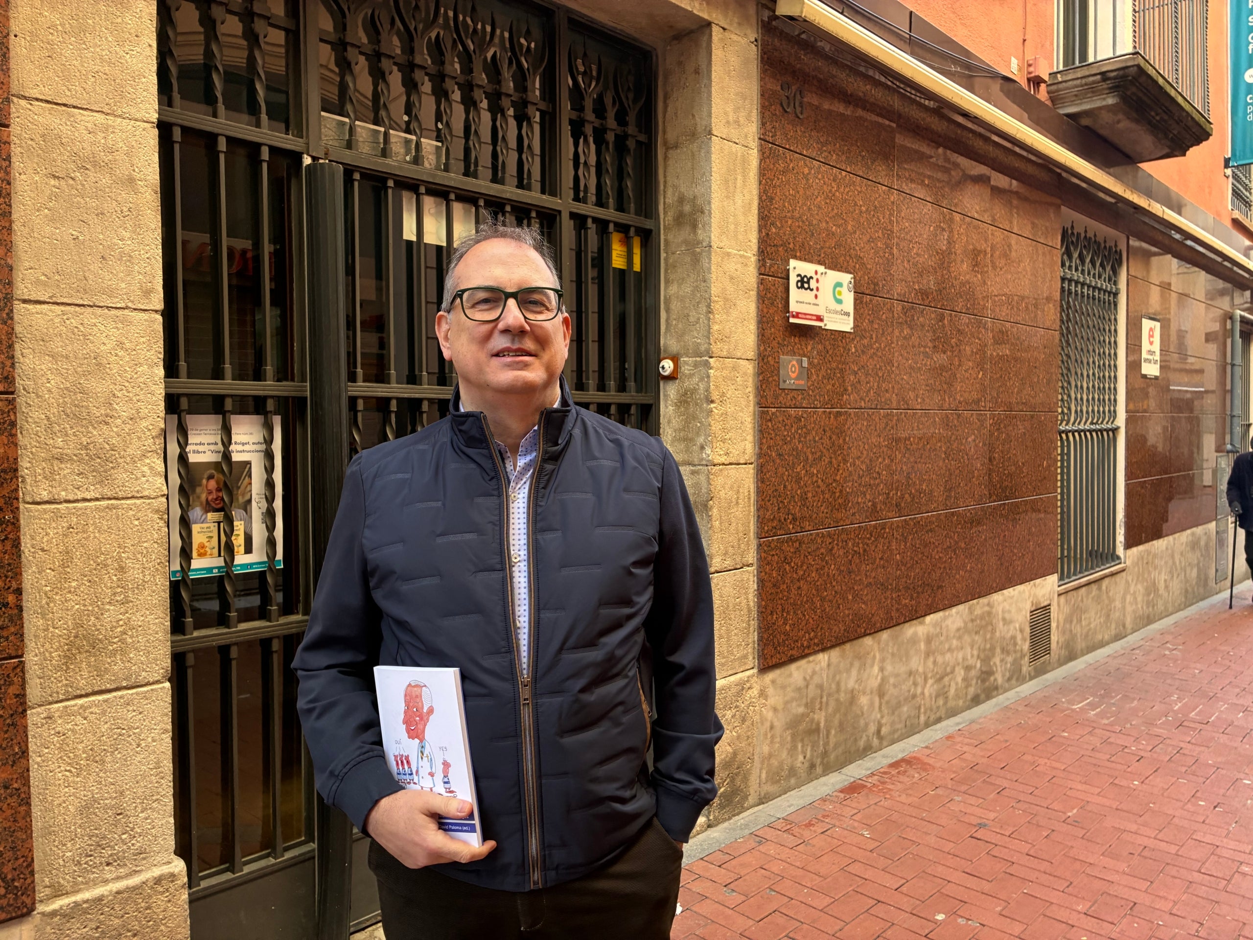 David Paloma, amb el llibre que ha coordinat sobre el seu pare davant del que va ser l'escola Cultura Pràctica al carrer Sant Pere de Terrassa | Vicenç Batalla
